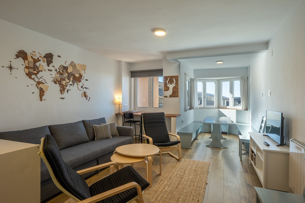 A modern living area features a comfortable gray sofa paired with two wooden armchairs. A round coffee table is positioned centrally on a natural fiber rug. A wooden world map adorned the wall, while large windows offer natural light and views of the surrounding area.