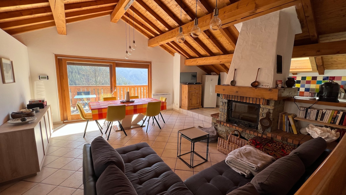 A spacious living area features a well-defined seating arrangement with a dark sofa and a stylish coffee table. A large dining table is set under the wooden beams, and a fireplace adds a rustic touch. Expansive windows provide natural light and showcase the mountain view.