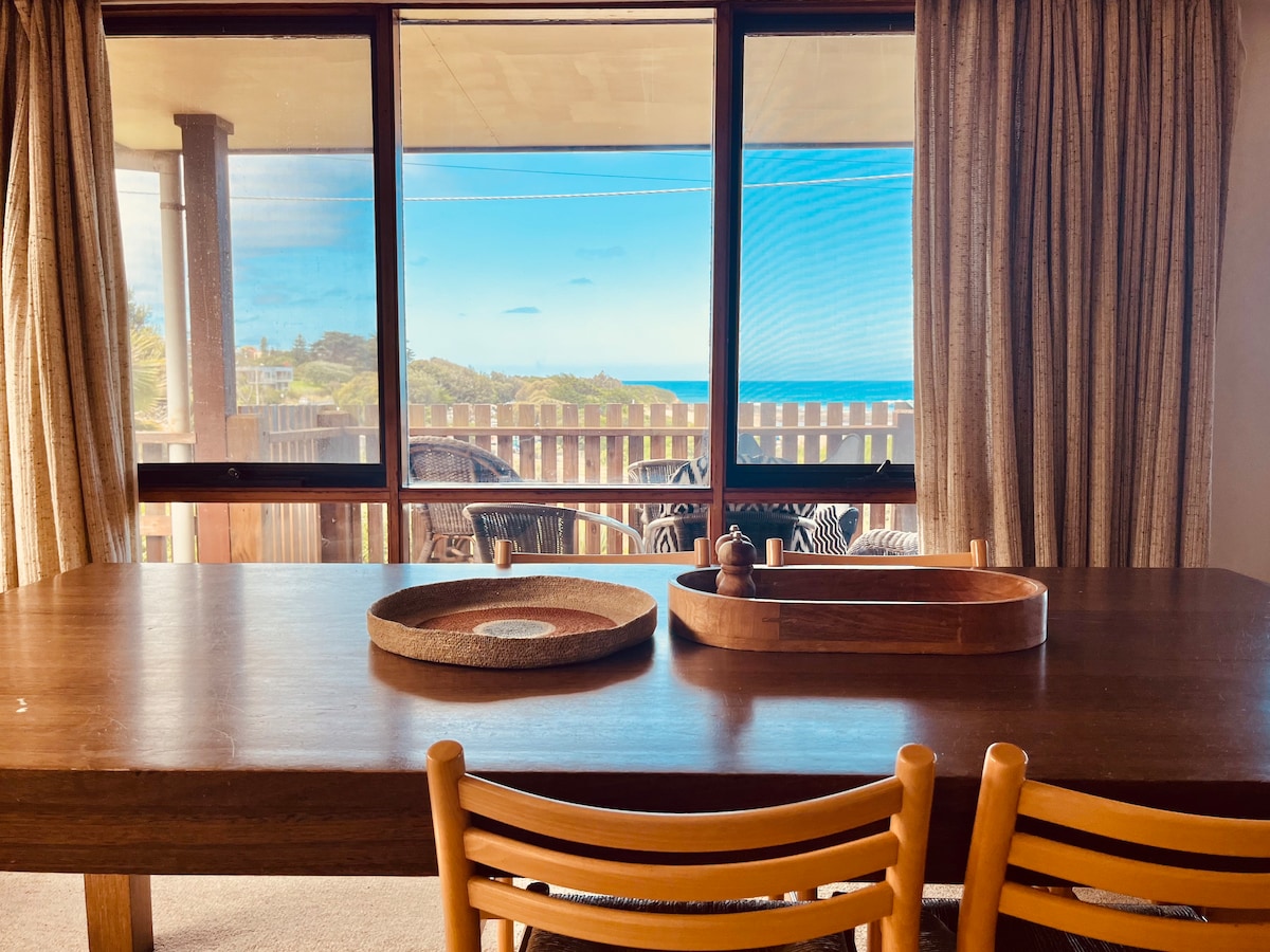 A dining area features a large wooden table, complemented by simple tableware. Natural light streams in through expansive windows, offering views of the ocean beyond. The space is framed by soft curtains, creating a warm and inviting atmosphere.