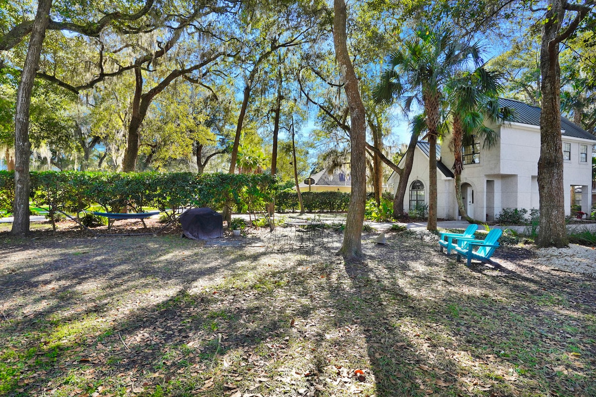 A serene outdoor area is framed by mature trees, providing shade and a natural ambiance. The space features a firepit, BBQ grill, and outdoor seating arrangements. A hammock is subtly positioned among the trees, inviting relaxation amidst the tranquil surroundings.