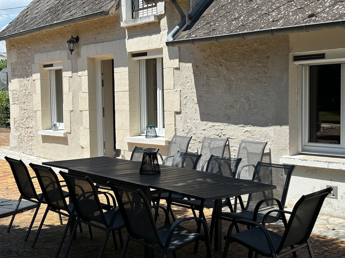 An outdoor dining area is shown, featuring a large grey table surrounded by numerous black chairs. The textured stone wall of the house is visible in the background, with large windows and a lantern enhancing the space. A calm atmosphere is observed in the well-lit area.