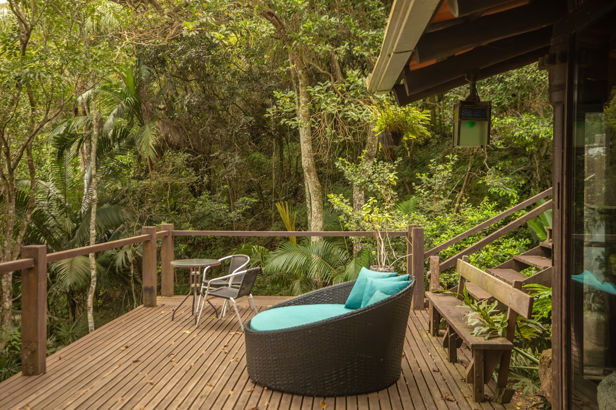 A wooden deck surrounded by lush greenery features a round wicker lounge chair with blue cushions. Nearby, a small metal chair is positioned for additional seating. The tranquil setting is enhanced by the sounds of nature, creating a peaceful retreat.