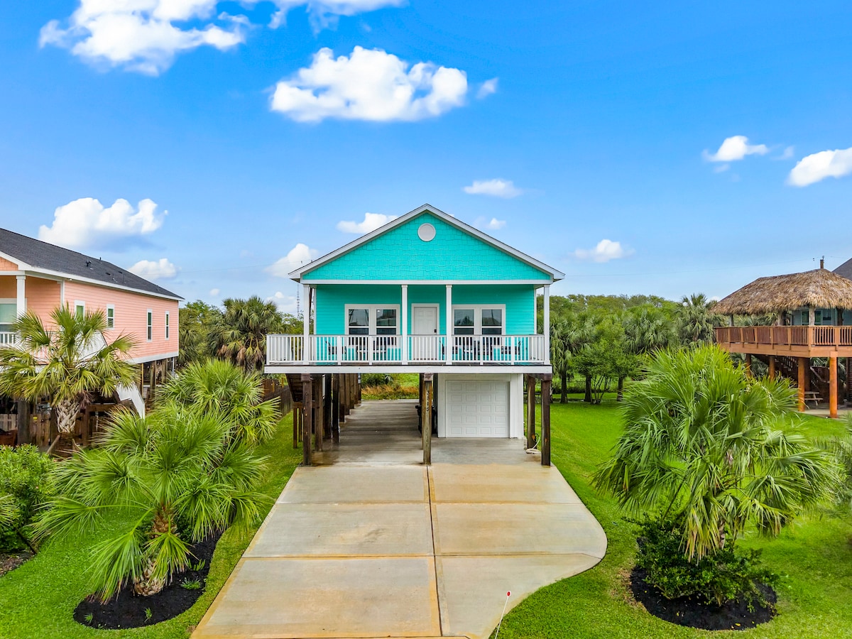 A vibrant turquoise home stands elevated on stilts, surrounded by lush green grass and palm trees. The spacious concrete driveway leads to the garage beneath the house, with a welcoming porch featuring white railings that offers serene views of the surrounding landscape.