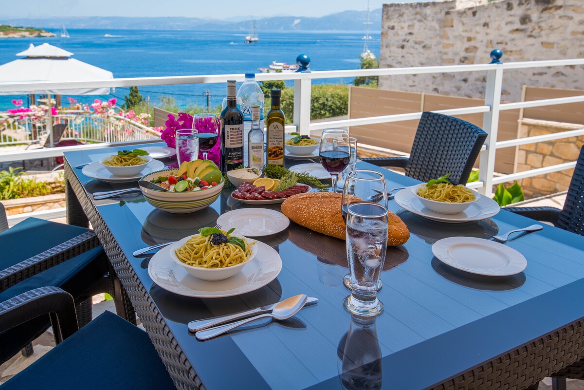 A dining table is set with plates of pasta, fresh salad, and a loaf of bread. Glasses of water and red wine are arranged neatly, with bottles of oil and vinegar nearby. The table overlooks the sea, featuring vibrant flowers in the background.