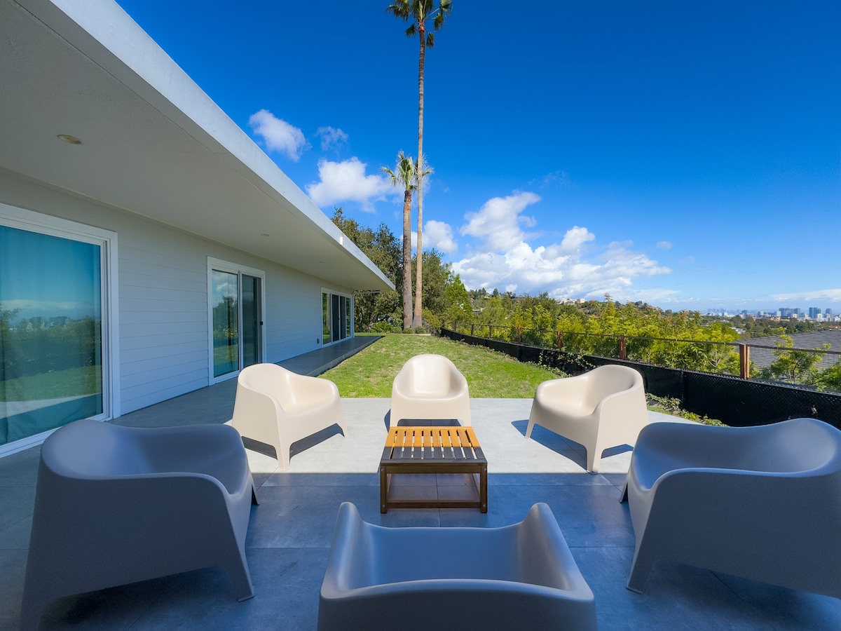 An outdoor seating area is arranged with several modern chairs surrounding a wooden coffee table. The space opens to a lush green backyard and showcases views of downtown Los Angeles under a clear blue sky, with a palm tree providing a touch of nature.