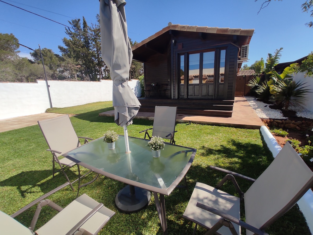 A peaceful garden area features a glass table surrounded by four chairs. A large umbrella provides shade overhead. In the background, the wooden house showcases a row of glass doors, offering a view of the serene outdoor space.