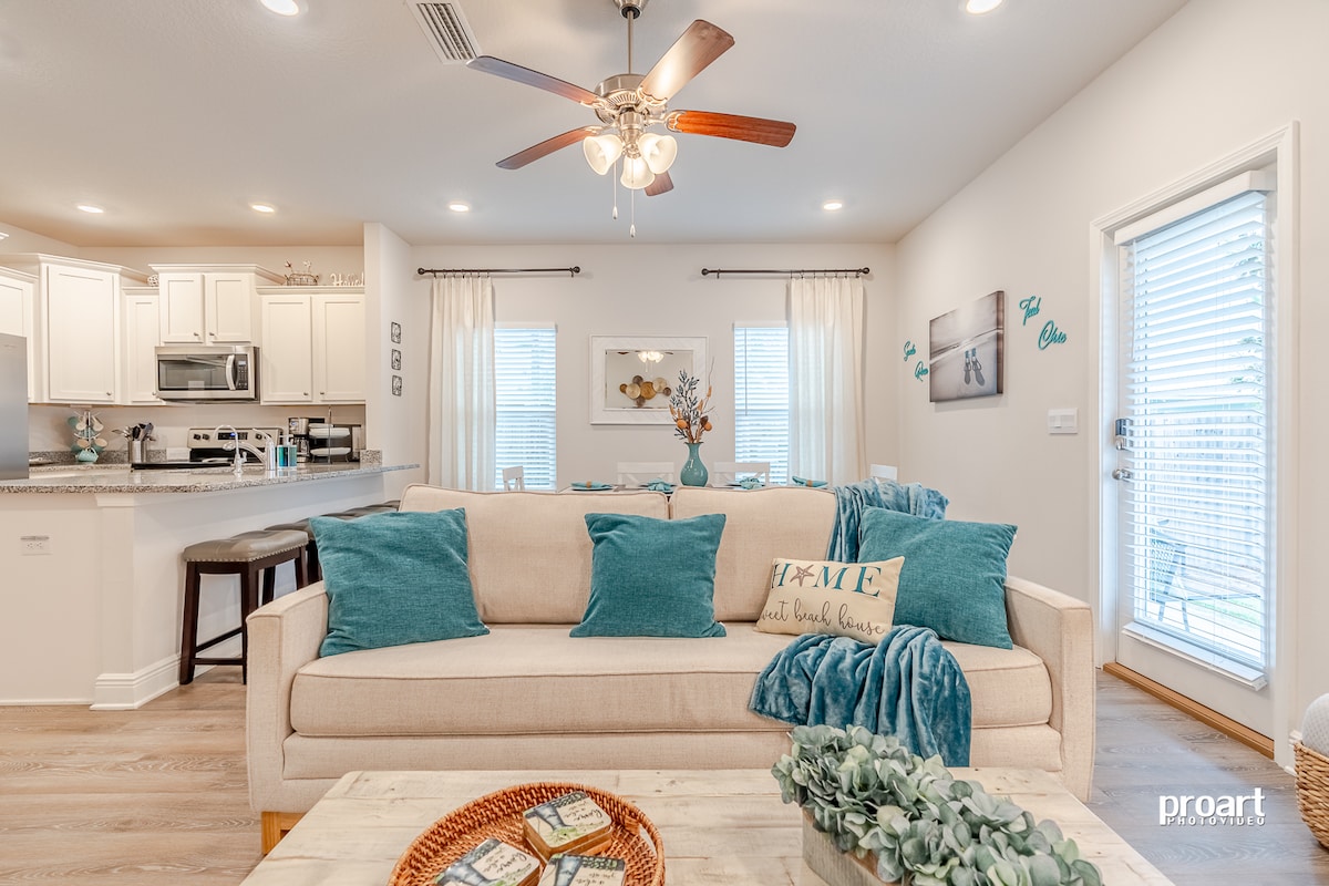 The living area features a light-colored sofa adorned with teal cushions, paired with a wooden coffee table. Natural light filters through large windows dressed with sheer curtains, illuminating a modern kitchen in the background equipped with white cabinetry and stainless steel appliances.