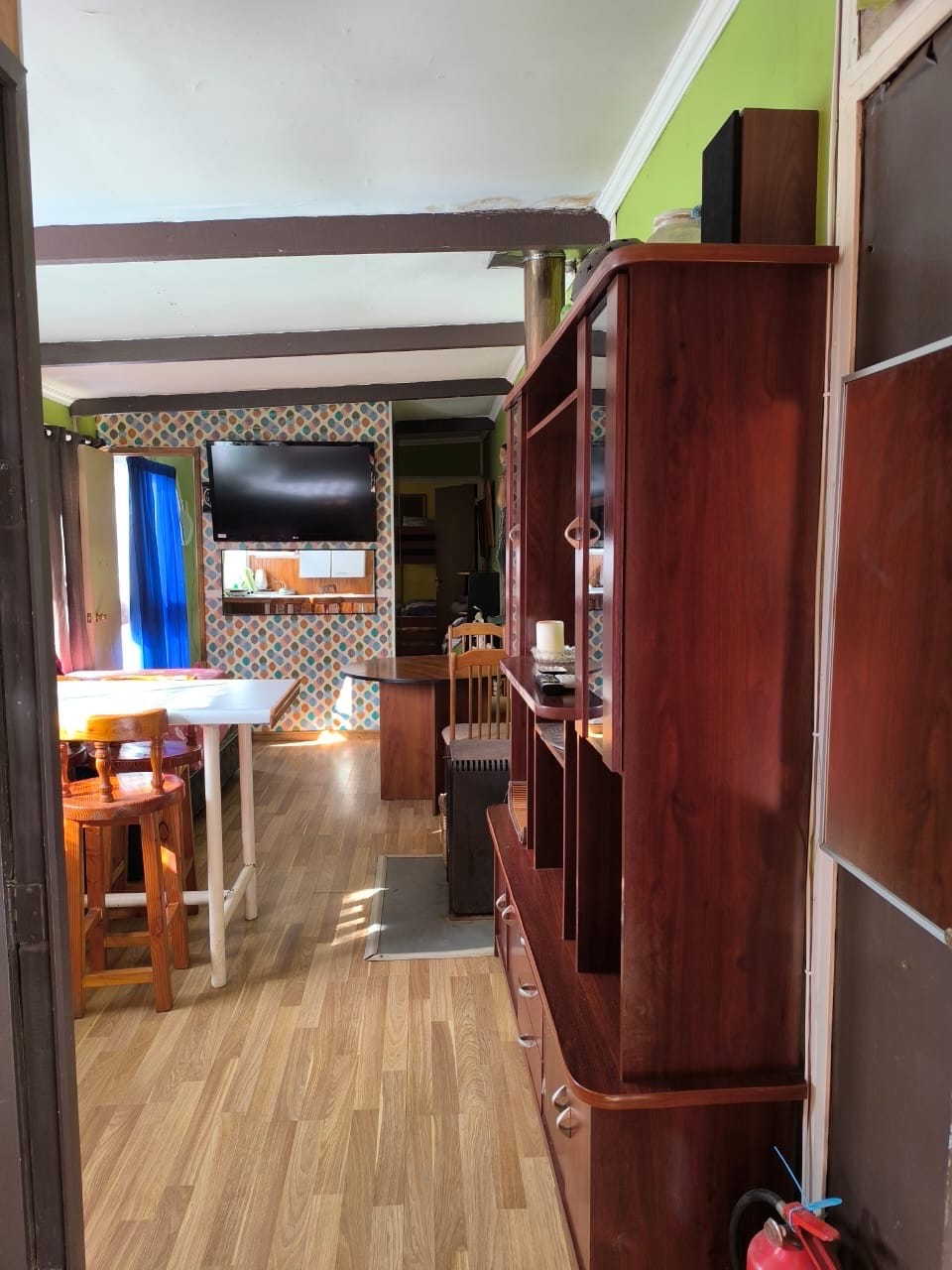 A view into a well-lit living space features wooden furniture, including a tall shelving unit and a dining table with chairs. A flat-screen television is mounted on a patterned wall, enhancing the cozy atmosphere. Light pours in from a blue curtain-draped window.
