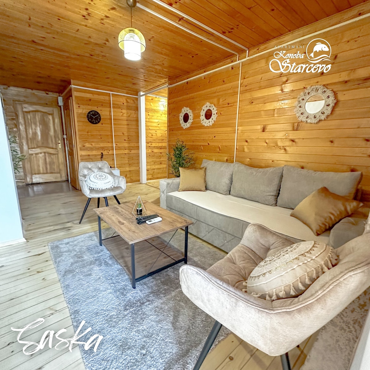 A welcoming living area features a comfortable sofa and two accent chairs surrounding a wooden coffee table. Wooden paneling adorns the walls, while a soft rug lies underfoot. Decorative wall hangings add subtle charm to the serene space.
