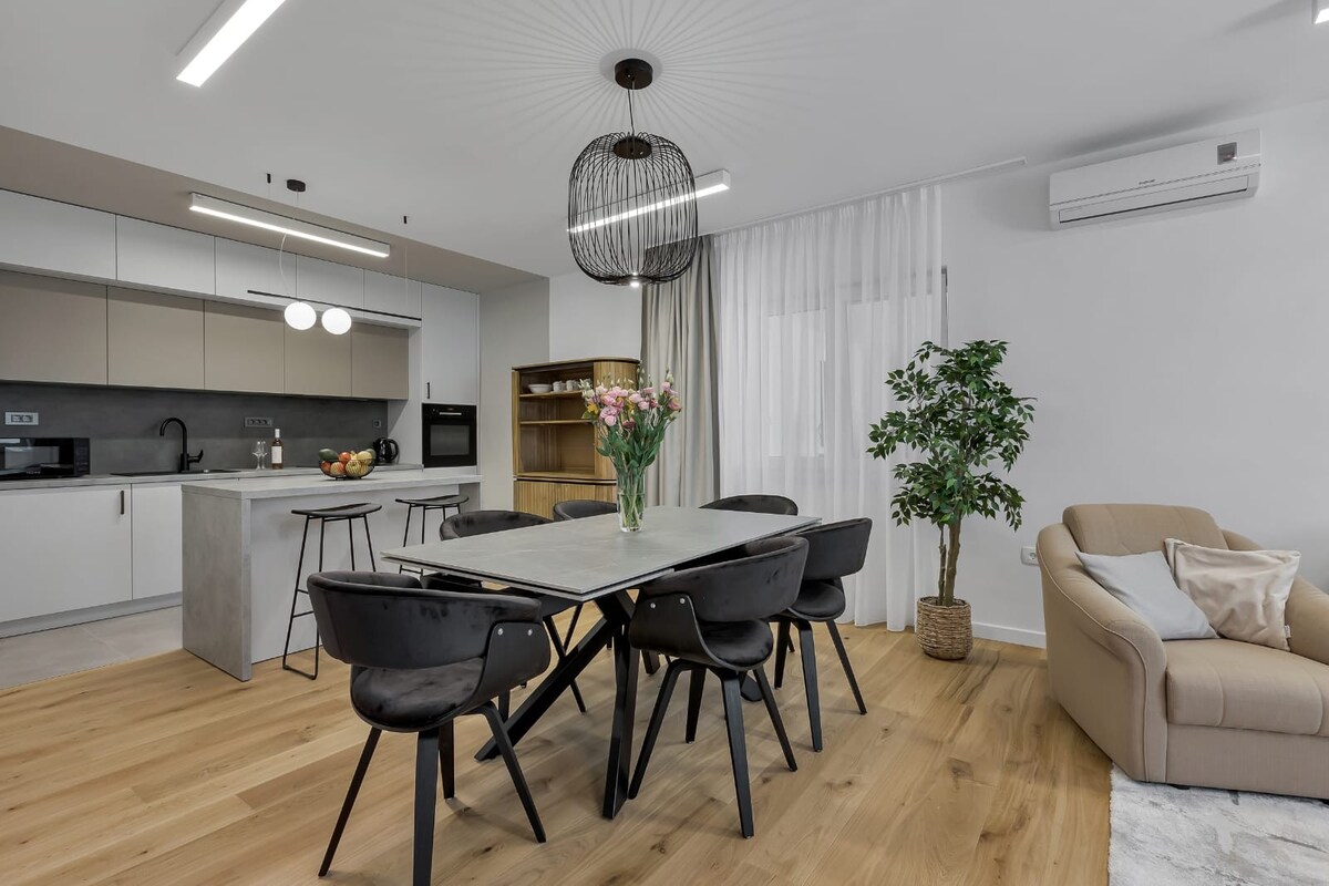 A modern dining area is presented with a sleek table surrounded by six black chairs. A unique pendant light hangs above, and natural light filters through sheer curtains. A potted plant adds a touch of greenery, while a comfortable sofa is located nearby.