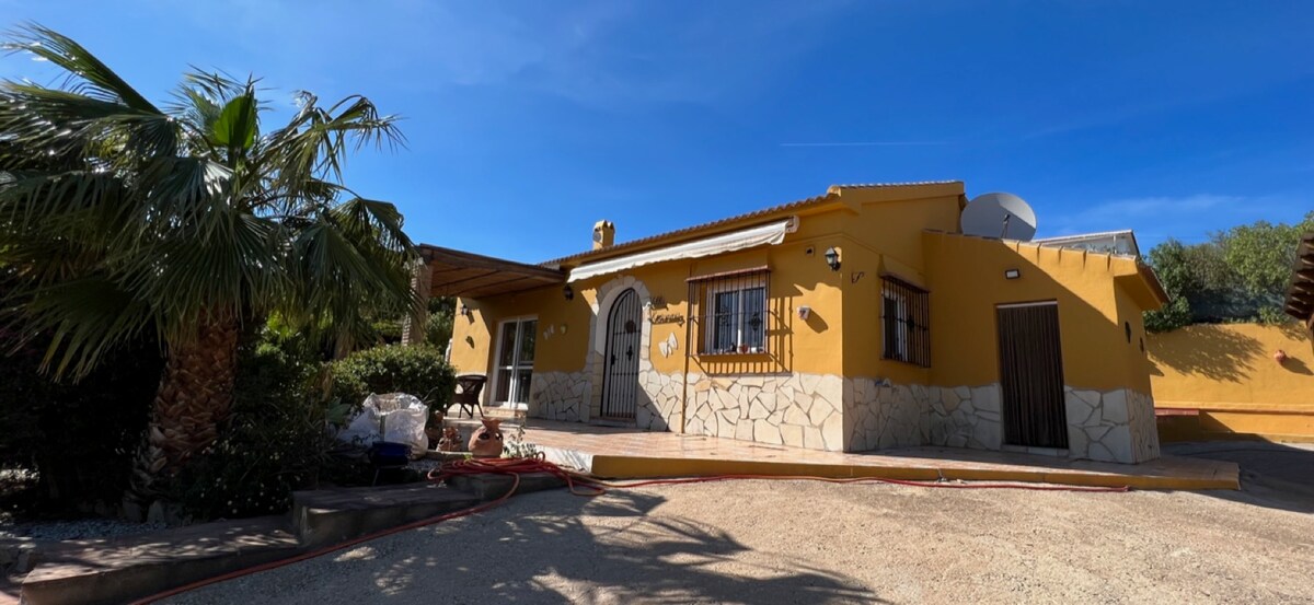 A single-story house with a warm yellow facade and stone accents is presented. The entrance features an arched door, flanked by windows. A covered porch with wooden beams offers shade, while tropical plants and a gravel driveway enhance the exterior ambiance.