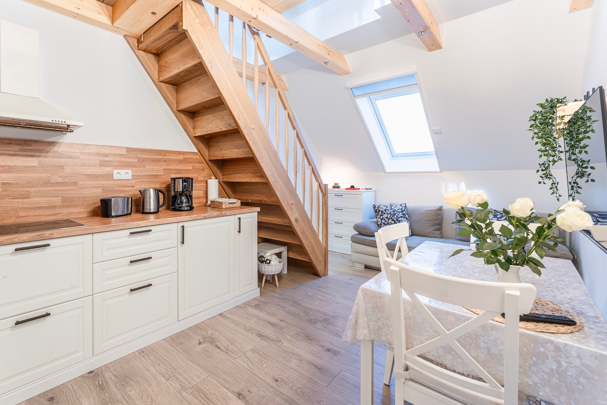 An open kitchen area features a wooden countertop and modern appliances, including a coffee maker and kettle. A staircase with wooden steps leads to the upper level. A cozy living space contains a comfortable couch and a small dining table adorned with flowers, enhancing the inviting atmosphere.