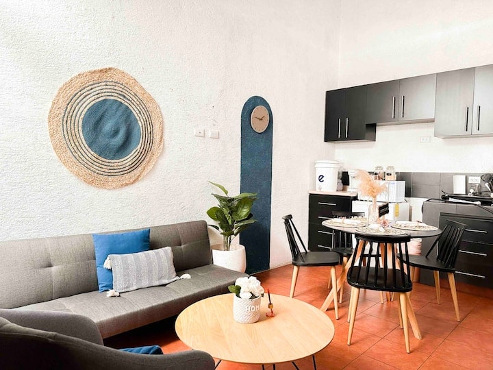 A modern living area features a gray sofa adorned with a blue accent pillow. A round wooden coffee table is positioned at the center. A dining area with a round table and black chairs is visible. The kitchen area includes dark cabinetry and appliances, complemented by natural decor.