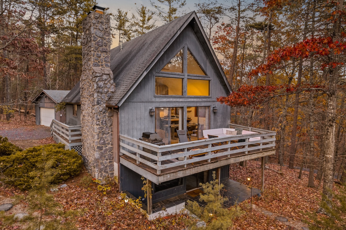 A charming cabin sits surrounded by autumn foliage, featuring a sturdy stone chimney and large windows that invite natural light. A spacious deck with seating areas offers a view of the wooded surroundings, while the exterior is clad in natural wood tones.