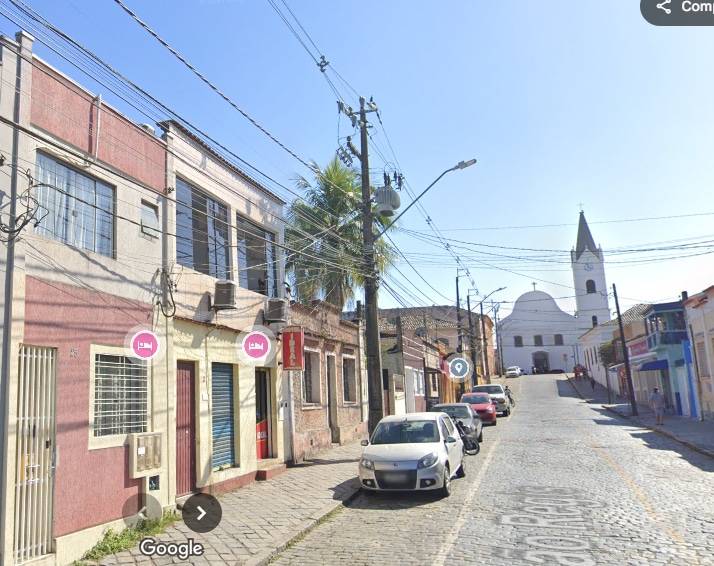 A street scene features two-story buildings lining a cobbled road, with a church visible at the end. Power lines run along the street, with a palm tree adding a natural element. A parked car is seen, and pedestrian pathways lead towards the church in the background.