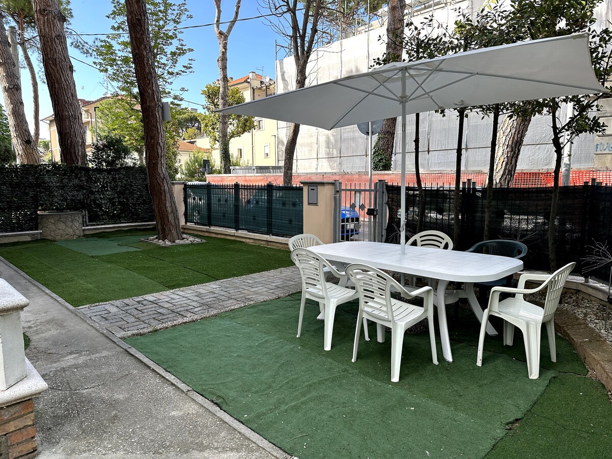 A spacious outdoor garden area is presented, featuring a large table surrounded by white chairs. A shade canopy provides protection from the sun, and meticulously maintained grass creates a comfortable setting. Tall trees line the perimeter, contributing to a serene environment.