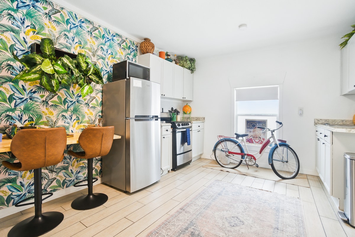 A bright kitchen area is complemented by a lively tropical-themed wallpaper. It features a stainless steel refrigerator and a gas stove surrounded by white cabinetry. Two brown leather bar stools are positioned at a small dining table. A bicycle is leaning against the wall, adding a casual touch.