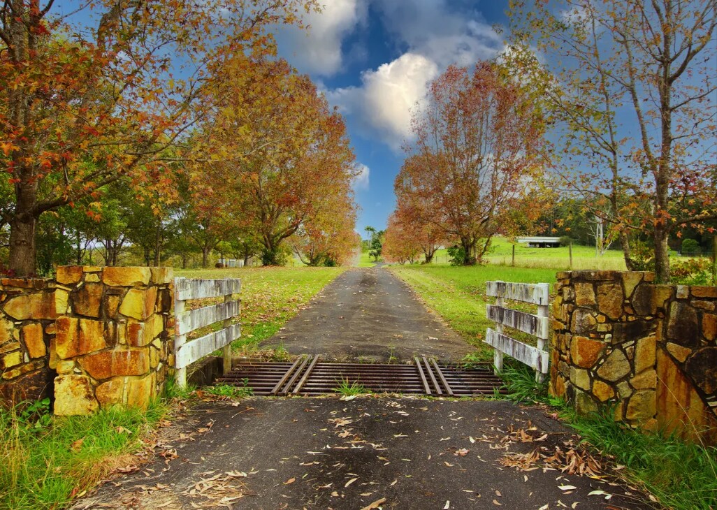 A gravel driveway stretches ahead, flanked by trees showcasing autumn foliage. Stone pillars mark the entrance, while a wooden gate rests open, inviting guests into the expansive countryside. Lush green grass surrounds the path, under a clear sky with scattered clouds.