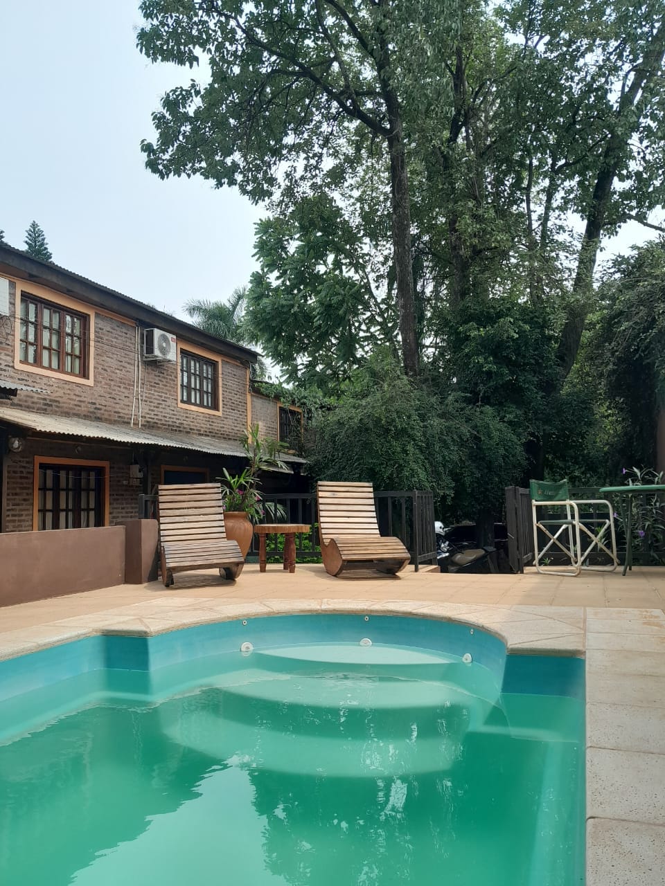A serene pool area is featured with clear water complementing the surrounding greenery. Two wooden loungers are positioned beside the pool, and a wooden table with chairs is visible in the background, along with a rustic two-story building set amid lush trees.
