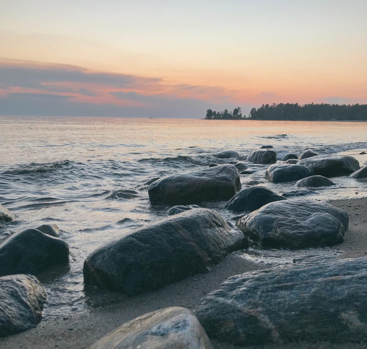 A serene shoreline is captured, featuring smooth stones scattered along the wet sand. Gentle waves kiss the rocks, while the horizon displays soft pastel hues from the setting sun, illuminating the tranquil waters of Georgian Bay.