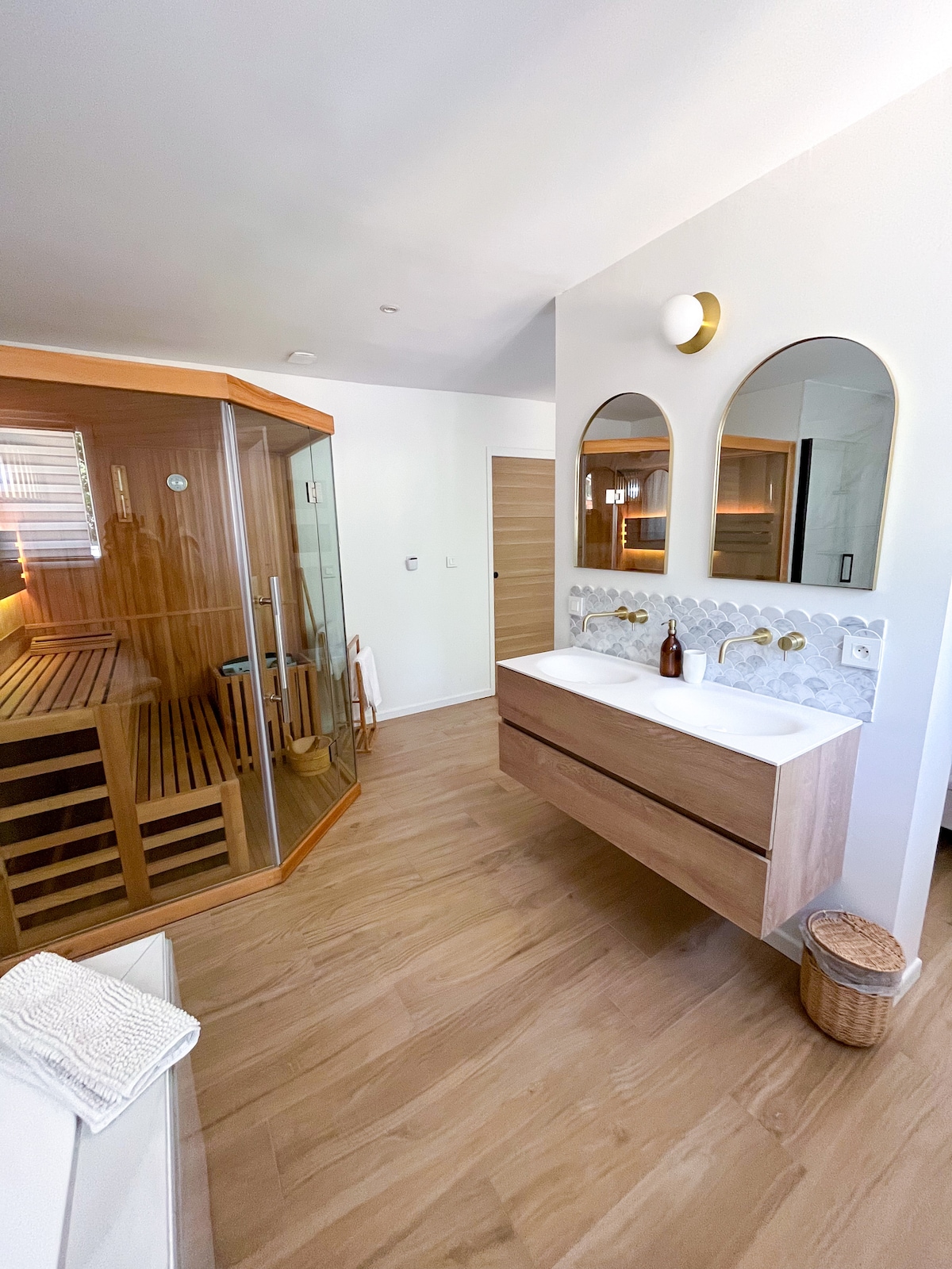 A spacious bathroom features a modern double vanity with two circular mirrors. A sauna is visible to the left, showcasing warm wooden slats. Light wood flooring enhances the bright atmosphere, while a woven basket and decorative elements add subtle detail.