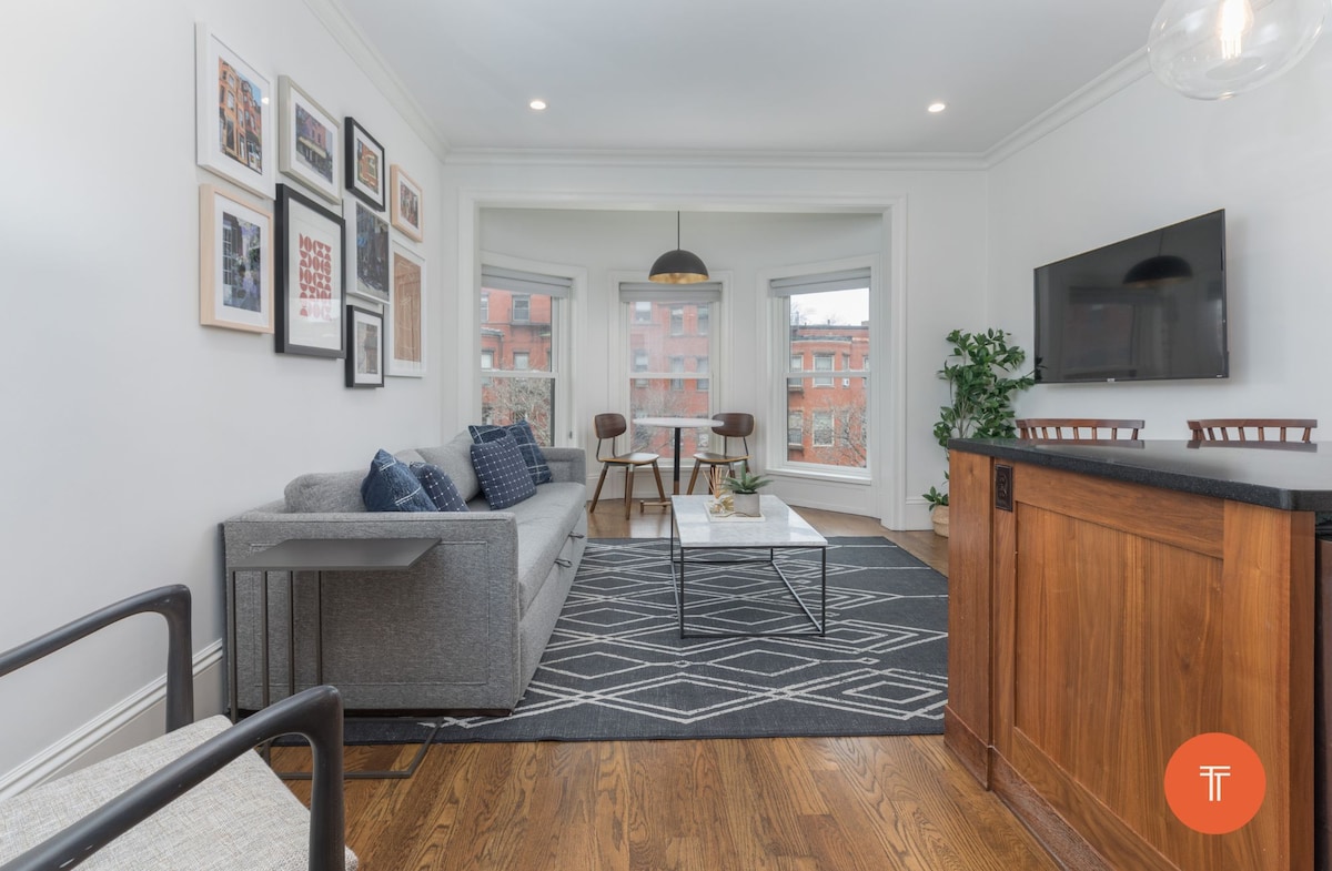A bright living area is equipped with a gray sofa and two patterned throw pillows. A minimalist coffee table sits on a geometric rug. Large windows allow for ample natural light, while a mounted television is positioned on the wall. Stylish bar stools complement the adjacent kitchen area.
