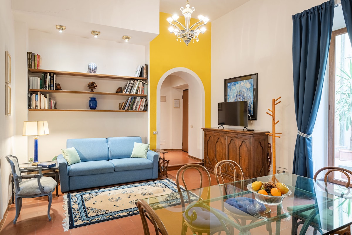 A living area is showcased with a light blue sofa adorned with soft pillows. A glass dining table is set with a bowl of fruit, while shelves display books and decorative items. A flat-screen TV is mounted on the wall, and natural light enters through the window with blue curtains.