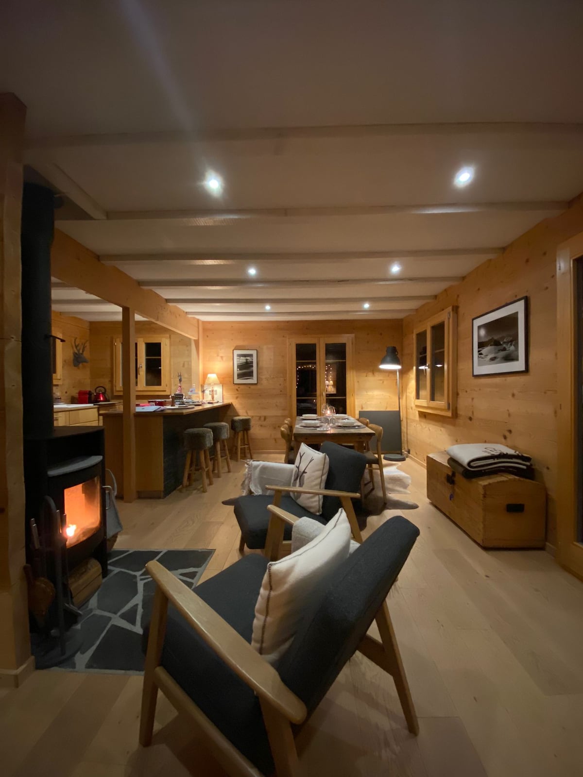 An open living space features wooden walls and ceiling beams, illuminated by recessed lighting. A cozy seating area includes two armchairs and a small table, while a wood stove provides warmth. The room is complemented by modern furnishings and a large window offering views of the surrounding nature.