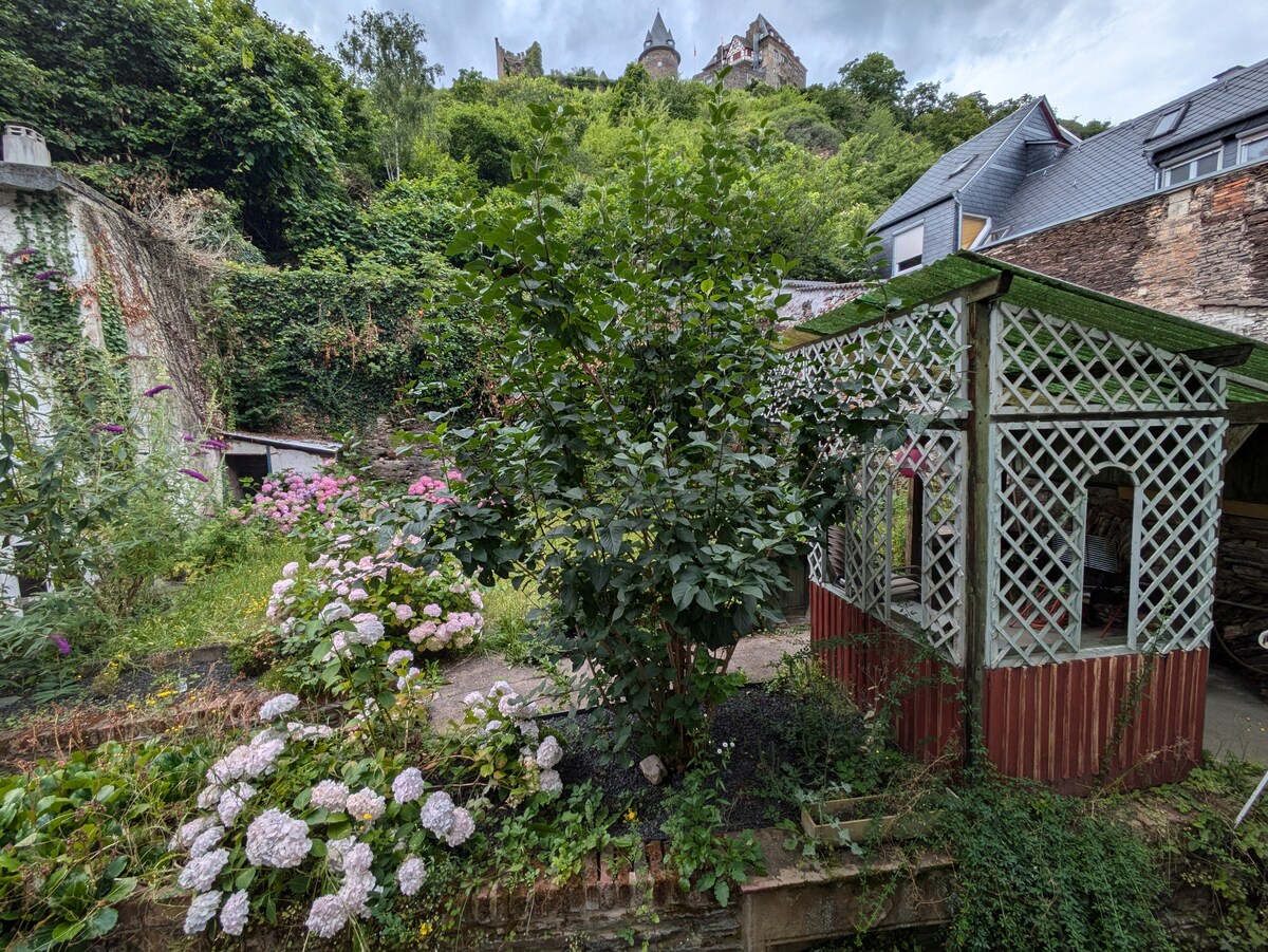 A vibrant garden features a green lattice gazebo surrounded by blooming hydrangeas and other flowers. The lush greenery on the hillside provides a scenic backdrop, with a castle perched above, creating an inviting atmosphere. A pathway leads through the garden, connecting to shaded areas.