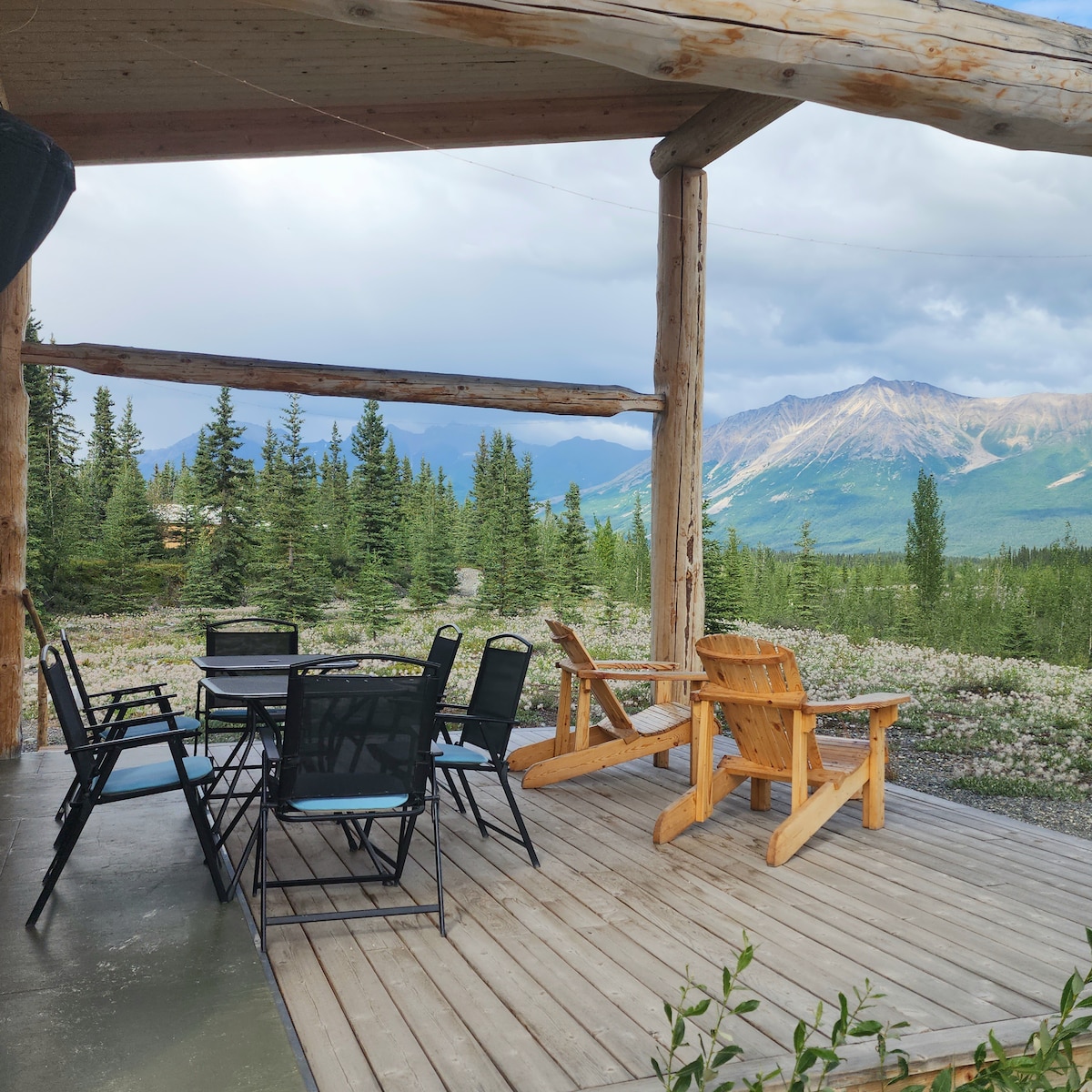 A spacious deck is furnished with wooden and metal seating, providing an inviting space to enjoy the outdoors. The backdrop features a panoramic view of distant mountains and lush greenery, under a partly cloudy sky.