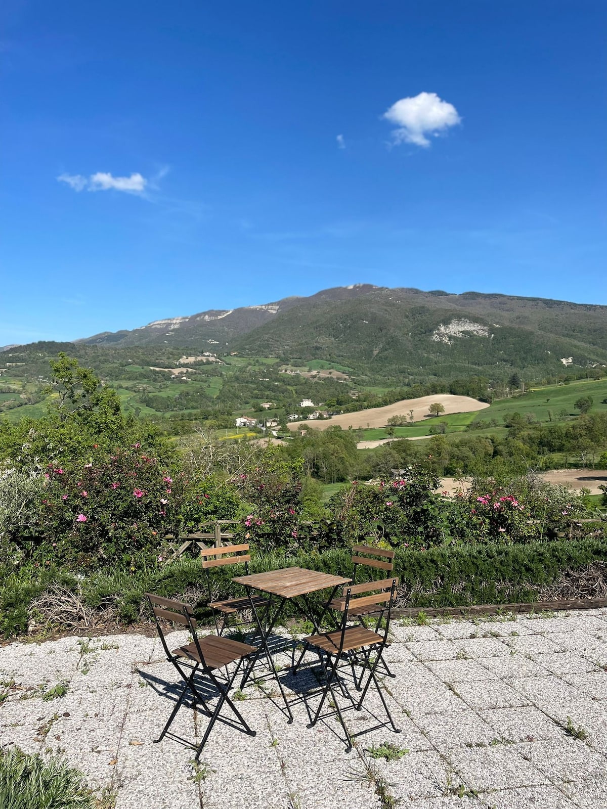 A small outdoor patio area is shown, featuring a simple table surrounded by two chairs. The backdrop reveals rolling green hills under a clear blue sky, with distant trees and buildings visible across the landscape.