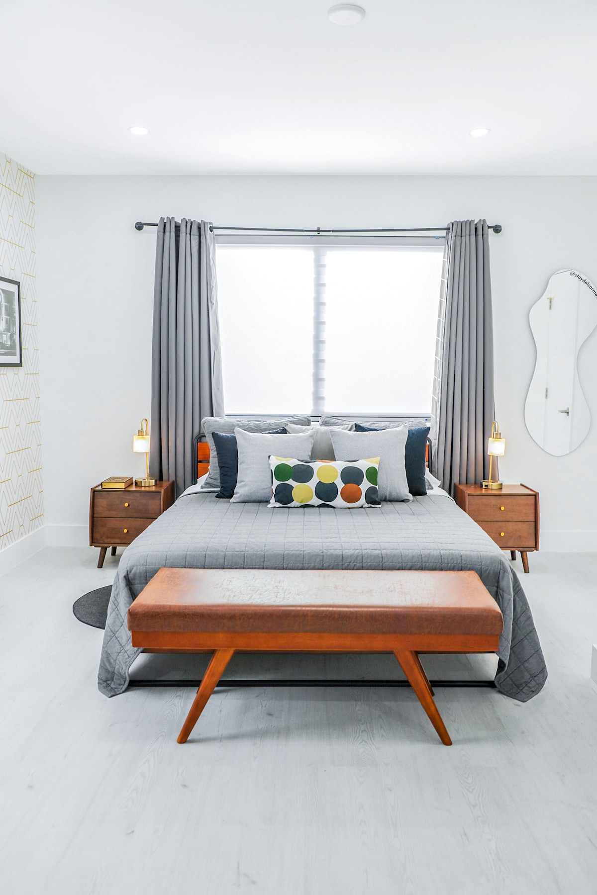 A modern studio bedroom is captured, featuring a comfortable queen bed adorned with patterned pillows. Two nightstands with lamps flank the bed, and a stylish leather bench sits at the foot. Large windows are fitted with sheer curtains, allowing soft natural light to fill the room.