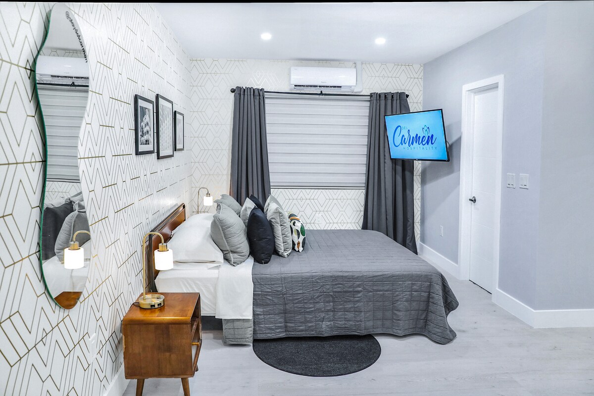 The well-furnished sleeping area includes a spacious queen bed dressed in soft gray linens and pillows. A flat-screen TV is mounted on the wall across from the bed. Natural light filters through the window, adorned with stylish gray curtains.