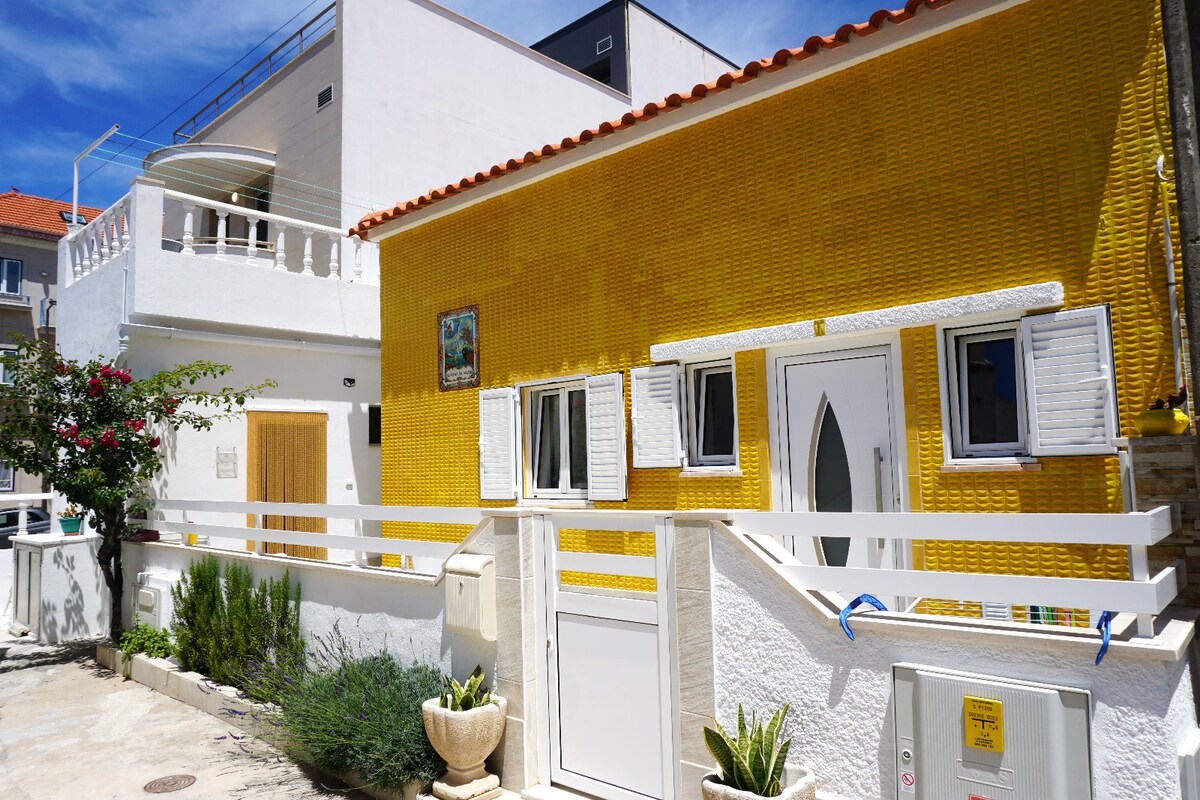 A brightly colored exterior of a small house is featured, showcasing a vibrant yellow façade and decorative white shutters. The entrance is highlighted by a uniquely shaped door. Surrounding greenery adds a touch of nature, while a terracotta roof peaks above the structure.