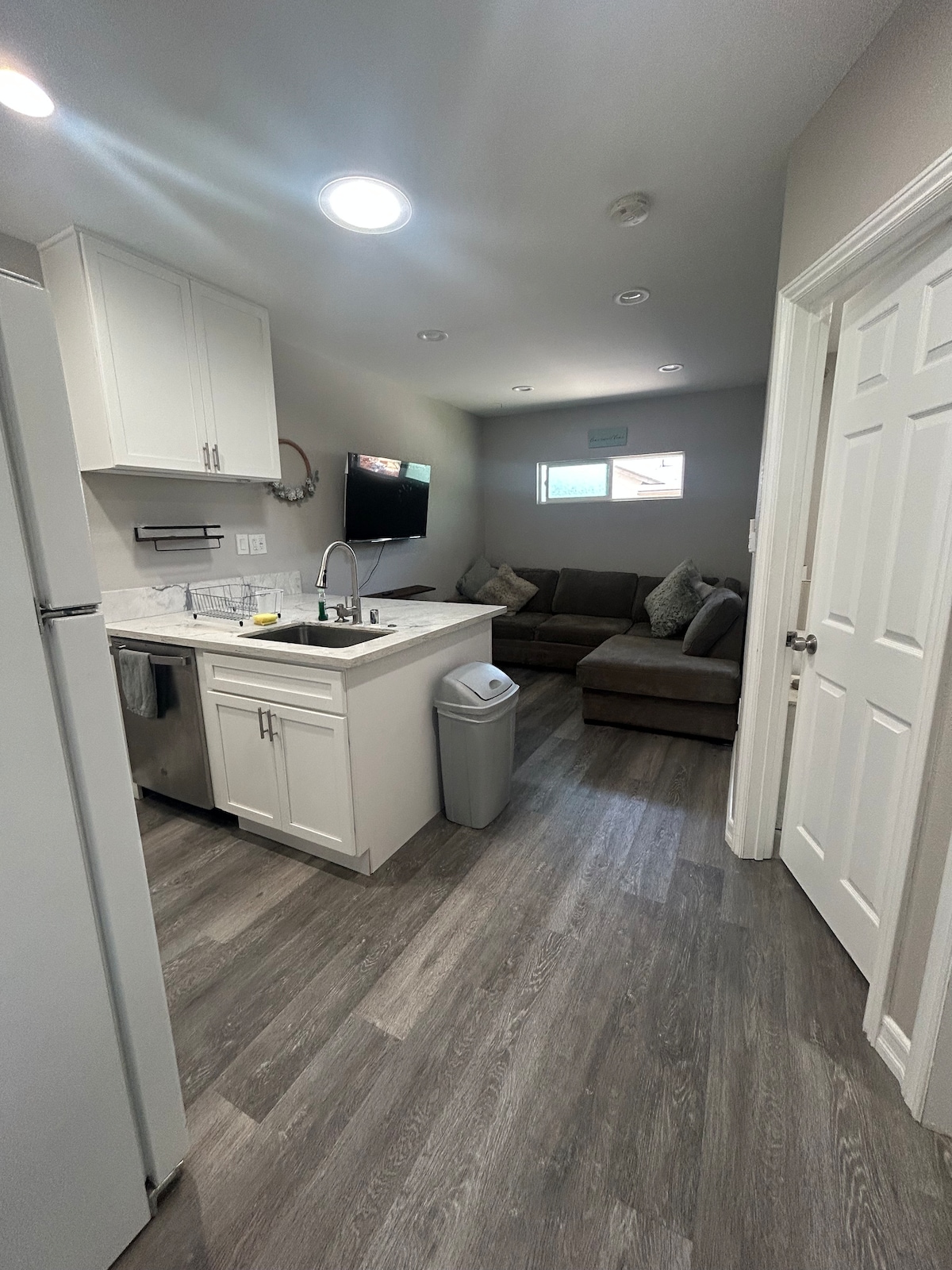 A compact living area is highlighted by a cozy corner sofa facing a wall-mounted television. An adjacent kitchen area features white cabinetry and a modern sink. Soft lighting is provided by recessed ceiling lights, enhancing the welcoming ambiance of the space.