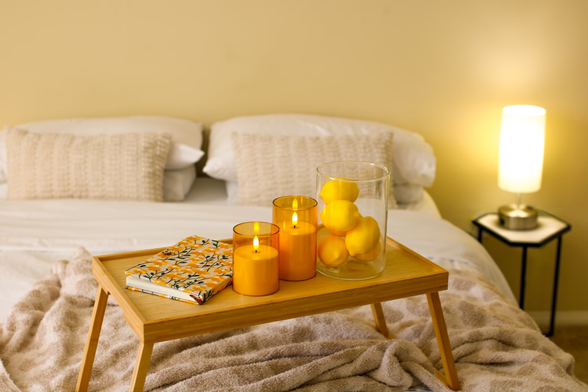 A spacious bed is presented with soft white linens, complemented by textured cushions. A wooden tray with decorative candles and a glass vase of lemons occupies the bed's surface, while a warm lamp provides gentle lighting nearby.