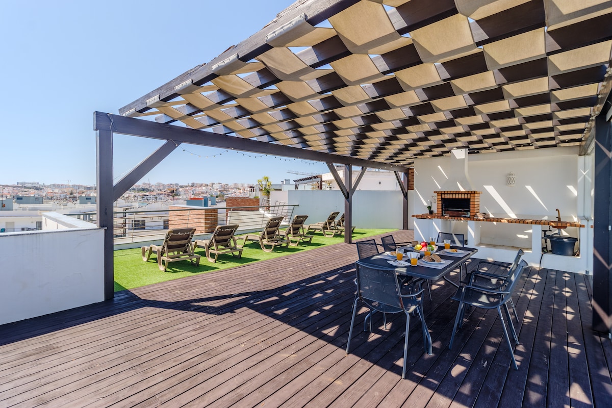 An expansive private roof terrace is featured, equipped with multiple lounge chairs and a dining area. A shaded pergola covers the dining table, which is set for a meal. The space is adorned with greenery, offering panoramic views of the surrounding area.
