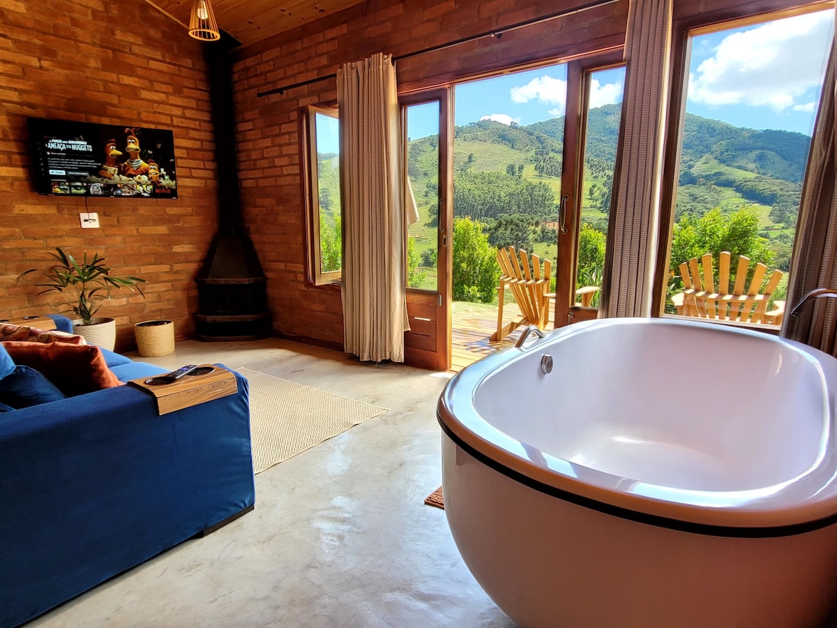 A spacious living area is highlighted by a large window offering panoramic mountain views. A freestanding bathtub is positioned near the window, while a cozy sofa and a smart TV are present, creating a comfortable environment with natural light and a peaceful ambiance.