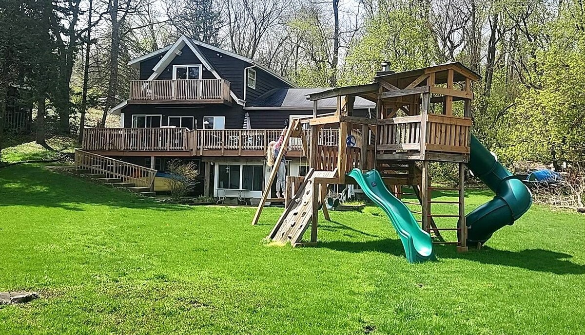 A spacious backyard is presented featuring a large wooden playset with slides and climbing features. The well-maintained green lawn surrounds a two-story lakefront house, showcasing a broad deck with ample space for outdoor seating and relaxation.