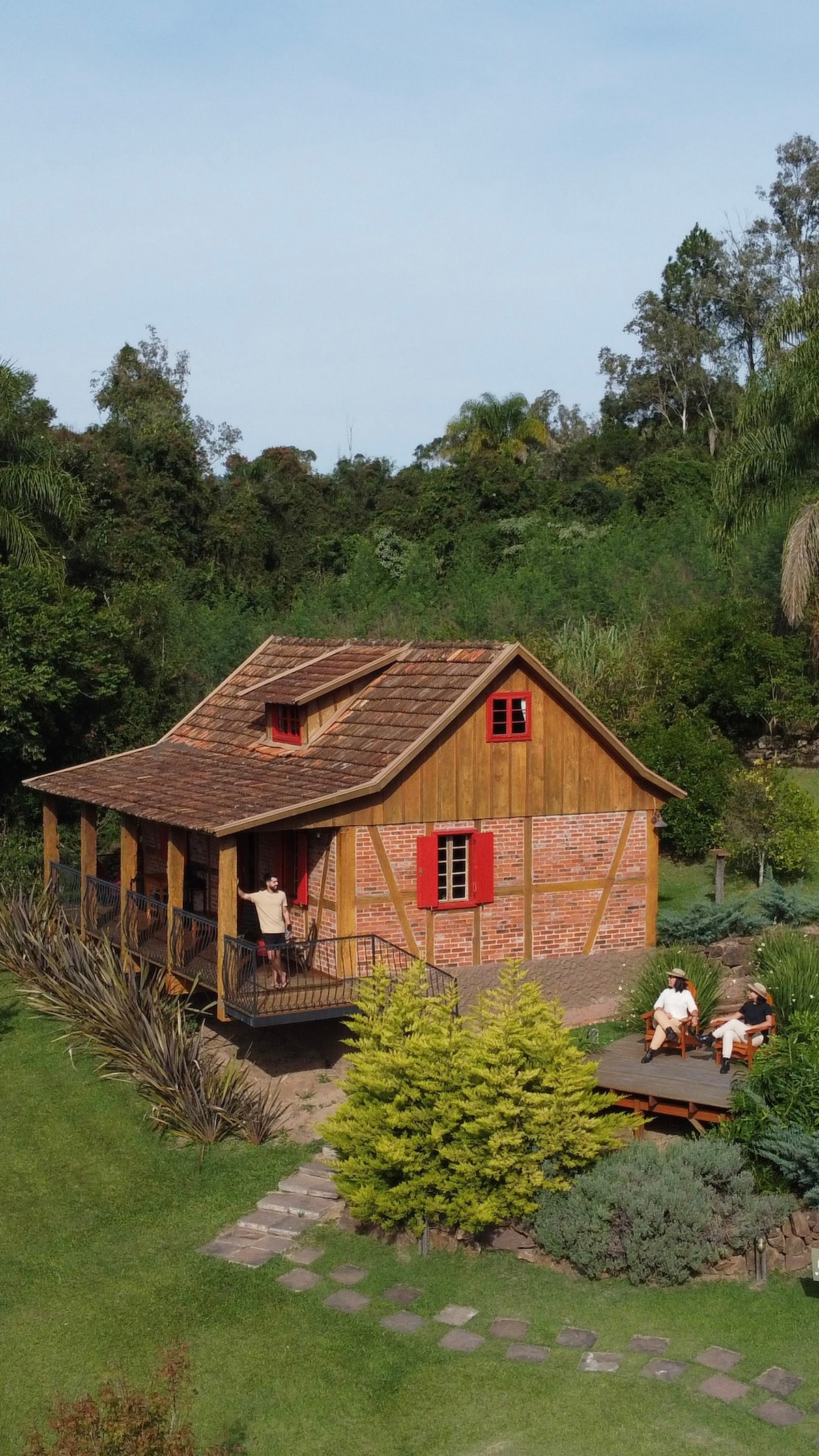 The red colonial chalet is surrounded by lush greenery, featuring a wooden structure with brick accents and characteristic red shutters. A spacious outdoor area includes seating arrangements, complementing the natural landscape, while pathways lead through vibrant plant life.
