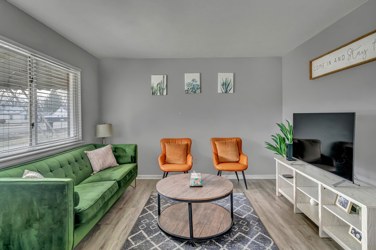 The living area features a green sofa accompanied by two orange armchairs, centered around a circular coffee table. A television sits on a white media console, and three framed plant artworks are displayed on the light gray wall. Natural light enters through two large windows with white blinds.