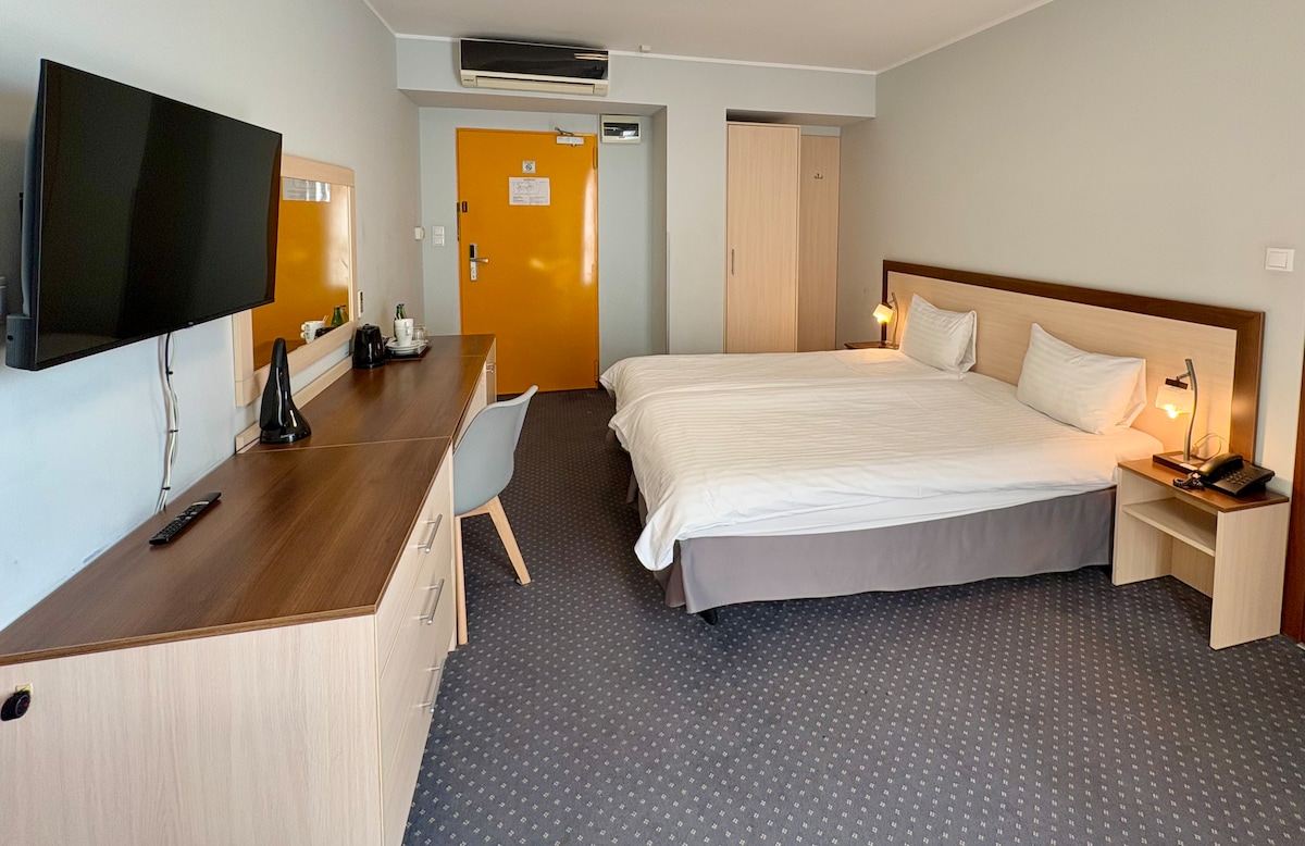 The room features a spacious bed with neutral bedding, complemented by bedside lamps for soft lighting. A television and work desk are positioned against one wall, while a wardrobe is visible nearby. The door displays a bright orange color, contrasting with the muted tones of the decor.