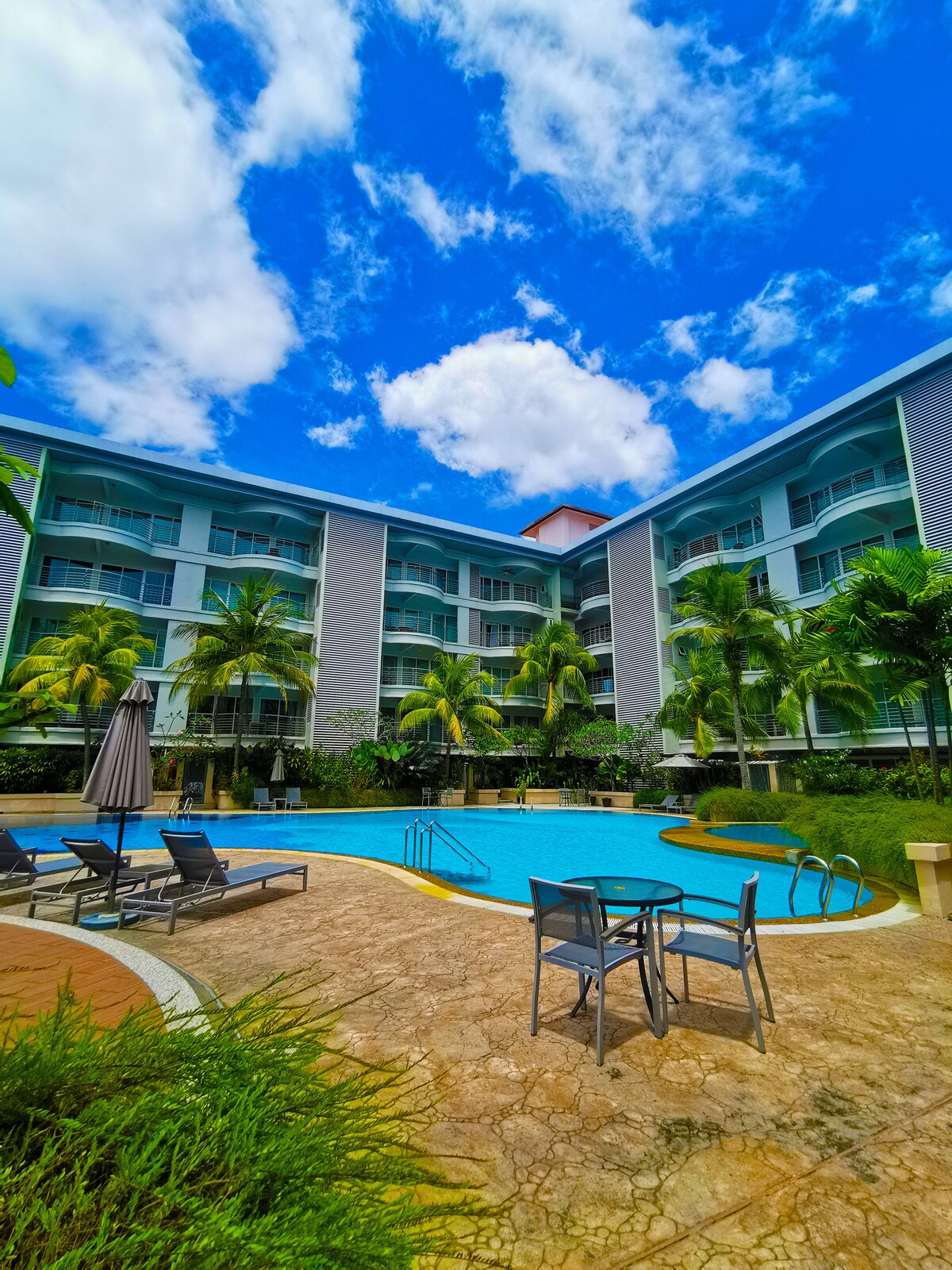 The image depicts a spacious outdoor pool surrounded by lush greenery and palm trees. Lounge chairs and a small table with chairs are positioned near the pool, under a bright blue sky with scattered clouds. The building stands in the background, showcasing multiple levels of balconies.