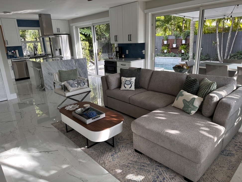 A spacious living area features a large sectional sofa adorned with decorative pillows, facing a contemporary coffee table. Wide glass doors invite natural light and provide views of the private pool and tropical landscaping beyond. A modern kitchen area is visible in the background.