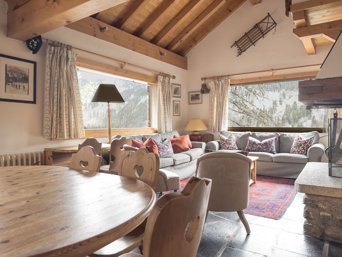 The sitting area features a spacious couch with decorative pillows, positioned near large windows offering views of the surrounding landscape. A wooden dining table with heart-shaped backs sits in the foreground. A traditional wooden beam ceiling adds warmth to the room.