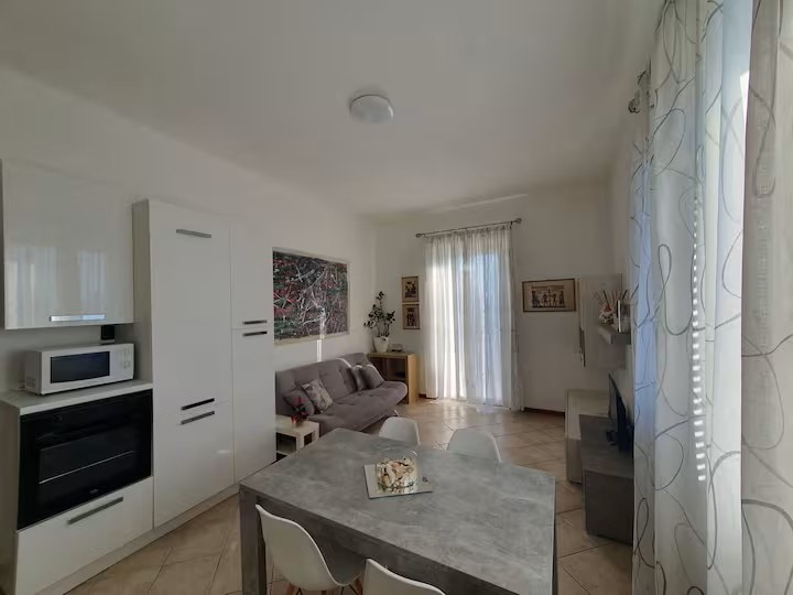 A modern living area features a grey sofa and a dining table with four chairs. Natural light streams through sheer curtains, highlighting the tiled floor and wall art. The kitchen area includes essential appliances, with a microwave and an oven integrated into cabinetry.