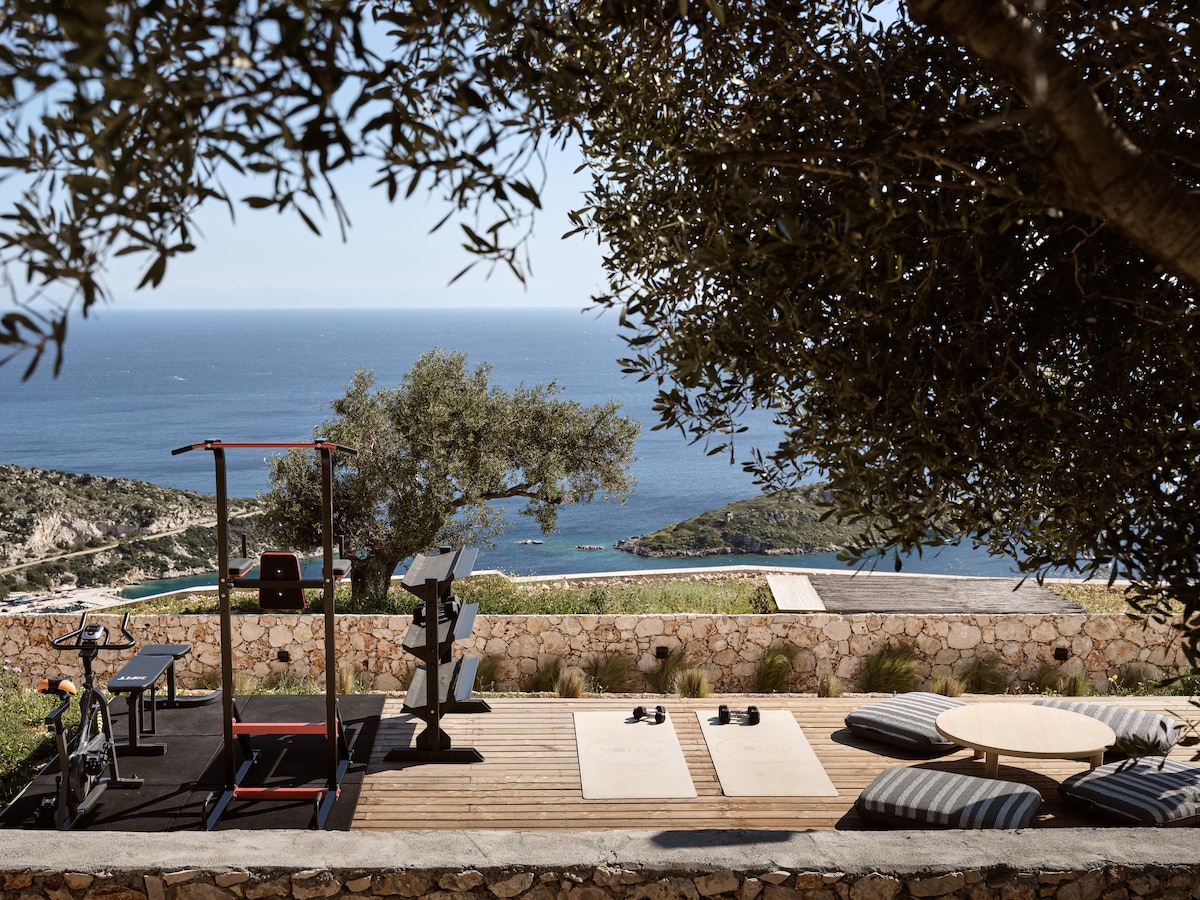 An outdoor fitness area features exercise equipment, including a stationary bike and weight station, overlooking the Ionian Sea. Two yoga mats are placed on a wooden deck, accompanied by a round table. Olive trees provide shade, enhancing the serene setting against the distant coastline.