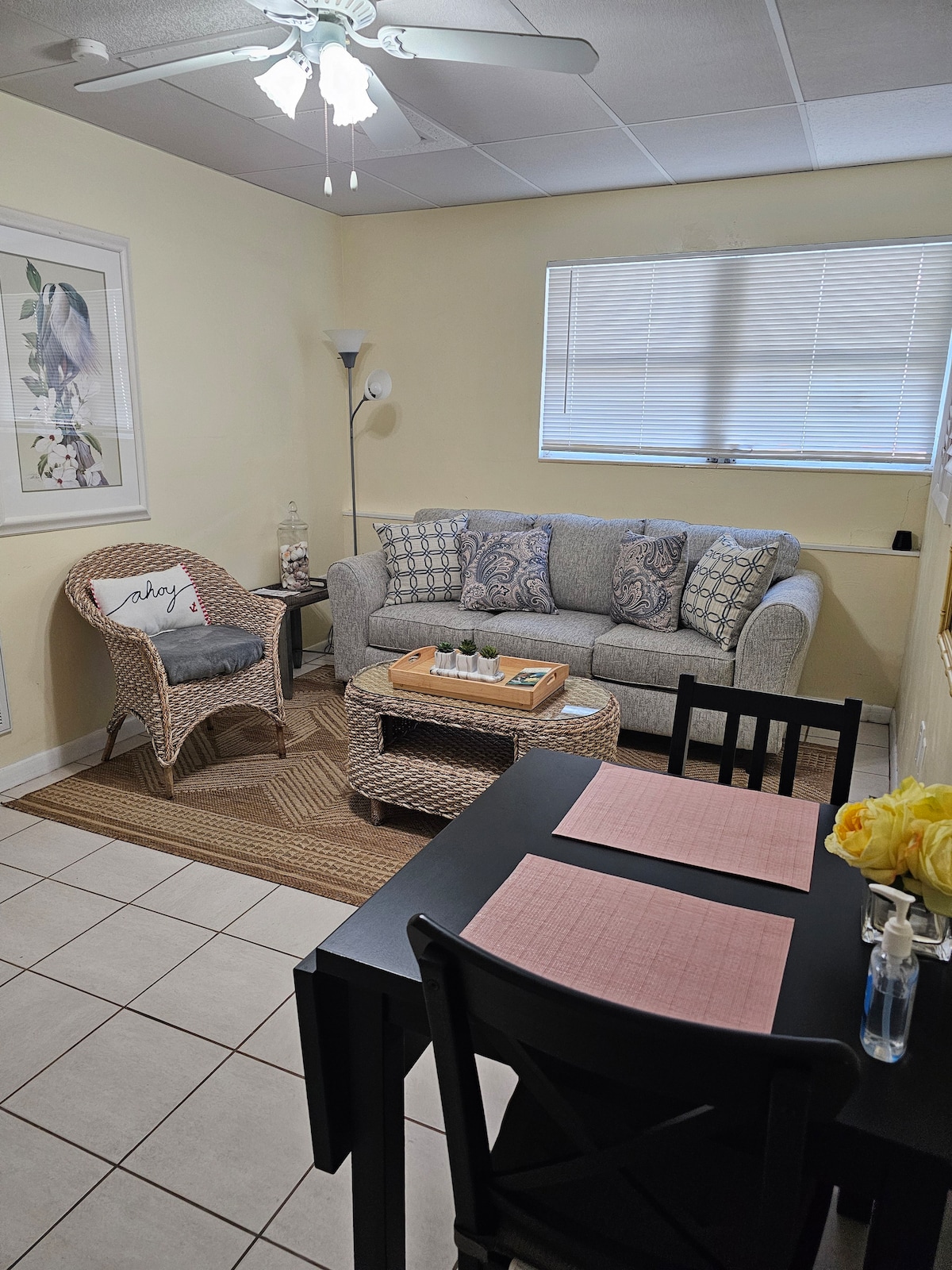 A cozy living area is highlighted by a light gray couch adorned with patterned pillows. A wicker chair sits nearby, and a small dining table is set with pink placemats. Natural light filters through a window, illuminating the tiled floor and decorative accents.