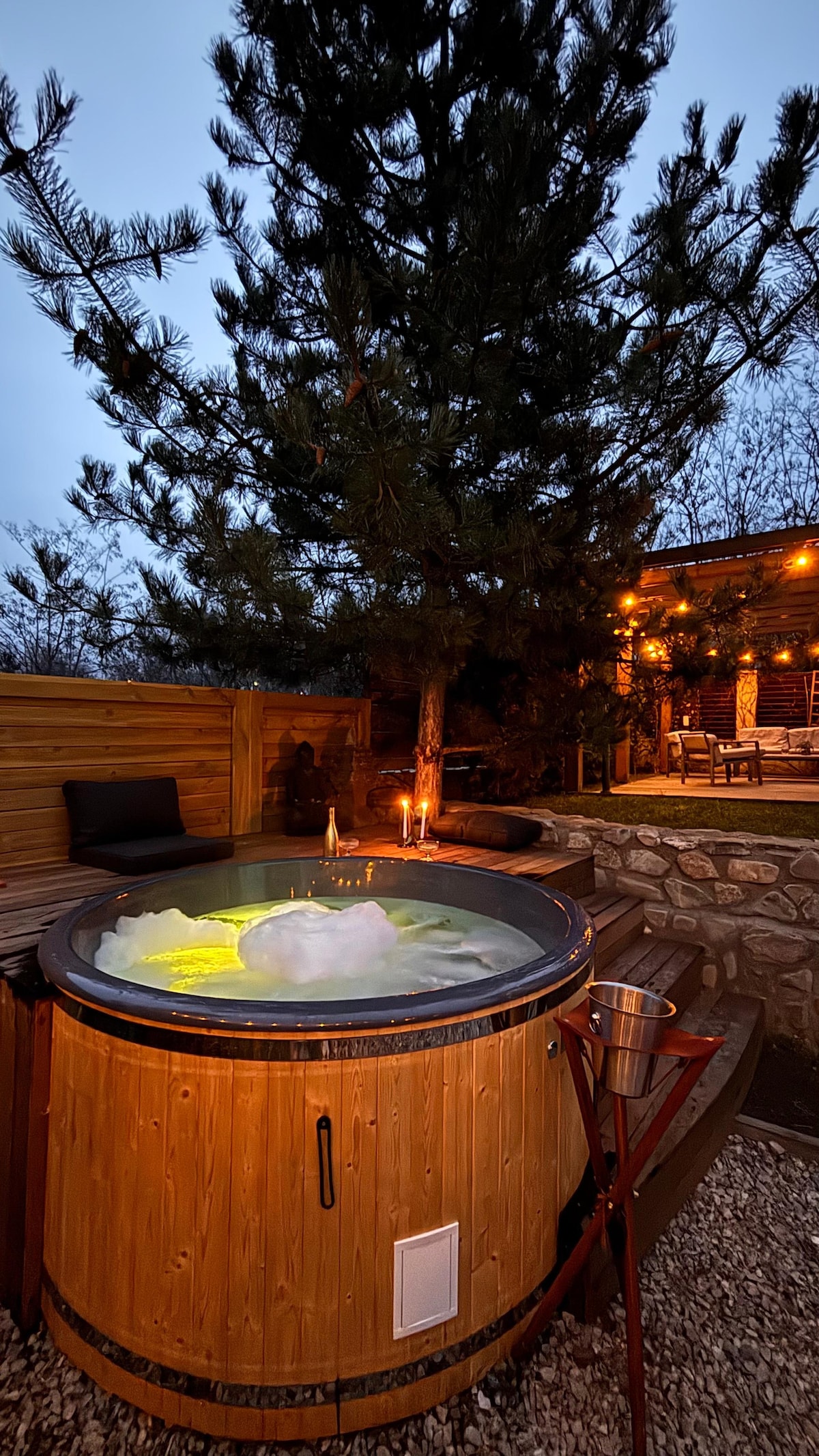 A wooden hot tub sits in an outdoor setting, surrounded by a stone wall and lush greenery. Candles are lit nearby, offering soft illumination. The area features comfortable seating and warm lighting from a building in the background, enhancing the serene ambiance.