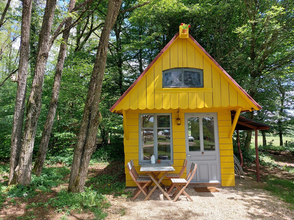 A small yellow house is set among trees, featuring large windows and a distinctive roofline. A small terrace with a wooden table and chairs is positioned outside, inviting relaxation. The surroundings exhibit a natural, lush atmosphere, enhancing a sense of seclusion.